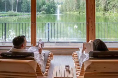 a man and woman sitting in chairs looking out a window at a waterfall at Hotel Lago della Creta in San Vigilio Di Marebbe