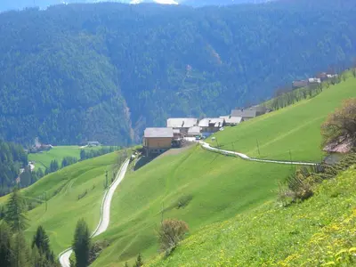 a green hillside with a farm and a winding road at Casa Gogn in San Vigilio Di Marebbe