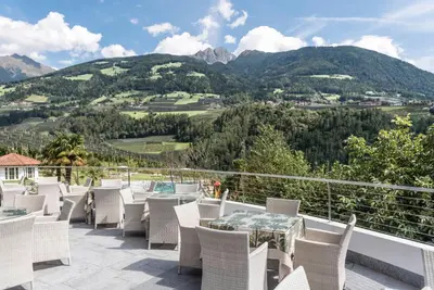 a patio with white chairs and tables and mountains at Hotel Hofbrunn in Rifiano