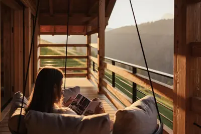 a woman sitting on a swing reading a book on a balcony at Lominus Dolomites Luxury Chalet for 2 in San Martino in Badia