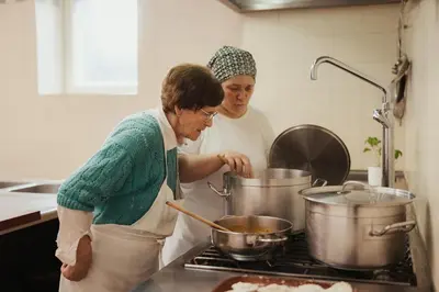 two women in a kitchen preparing food in pots at Pension Torr in San Martino in Badia