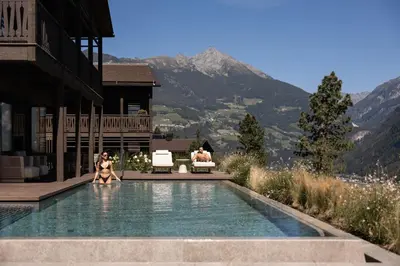 two women sitting in a swimming pool with mountains in the background at AMONTI Chalets in Molini di Tures