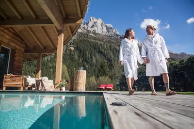 two women in white robes walking on a deck next to a swimming pool at Olympic SPA Hotel - Adults Only in Vigo di Fassa