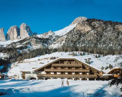 two people standing in the snow in front of a ski lodge at Hotel Rubino Deluxe in Campitello di Fassa