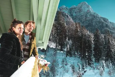 a man and woman standing next to a house in the snow at Hotel Erica in Tesero
