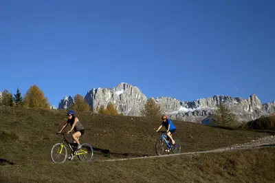 two people riding bikes on a hill with mountains in the background at Hotel Piné in Tires