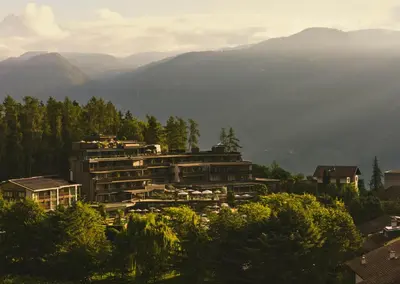 a building on a hill with mountains in the background at ALPIANA - green luxury Dolce Vita Hotel in Vollan