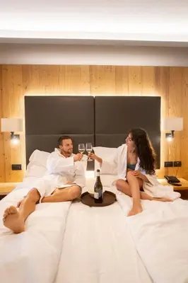 a man and a woman sitting on a bed with wine glasses at Hotel La Montanara Predazzo in Predazzo