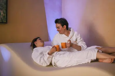 a man and a woman sitting on a bed with a cup at Hotel Arnika in Pozza di Fassa