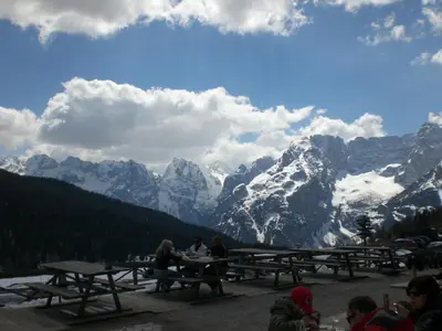 a group of people sitting at picnic tables in front of mountains at Hotel Villa Ines in Borca di Cadore