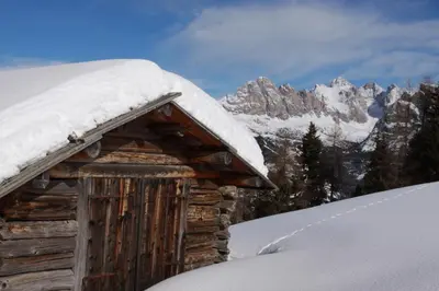 a log cabin covered in snow with mountains in the background at Pensione Vittoria in Colfosco