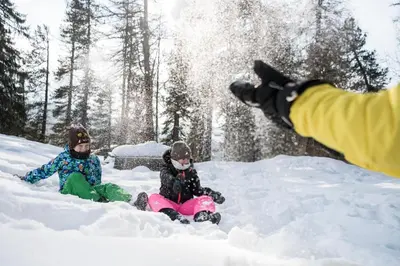 two children are sitting in the snow with a person jumping at Familienhotel Maria in Obereggen