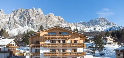 a large wooden building with snow covered mountains in the background at Camina Suite and Spa in Cortina dʼAmpezzo