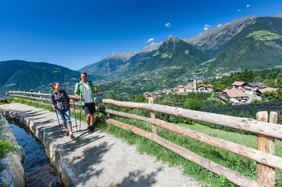 a man and a woman walking on a bridge with mountains in the background at Hotel-Garni Appartement Gartenheim in Schenna