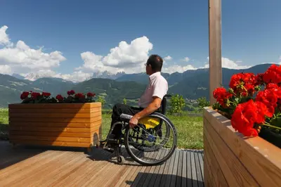 a man in a wheelchair sitting on a deck with flowers at Oberhauser Hof - Apartments in Feldthurns