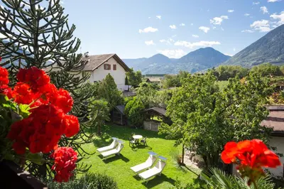 a garden with white chairs and red flowers and mountains at Hotel Gruberhof in Merano