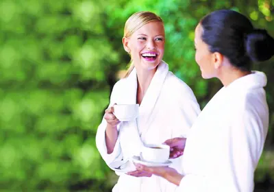 two women in white robes talking and holding coffee cups at Hotel Pienzenau Am Schlosspark in Merano