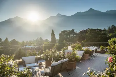 a deck with tables and chairs with mountains in the background at Hotel Pienzenau Am Schlosspark in Merano