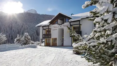 a building in the snow with the sun in the background at Residence Villa al Sole in Selva di Val Gardena