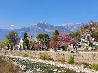 a stone wall next to a river with mountains in the background at Meranosuites - Therme in Merano