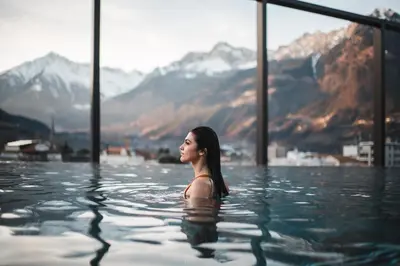 a woman is sitting in a swimming pool with mountains at Hotel Therme Meran - Terme Merano in Merano